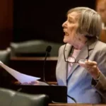 Older woman in a gray suit speaks at a podium with a microphone, holding papers and glasses.