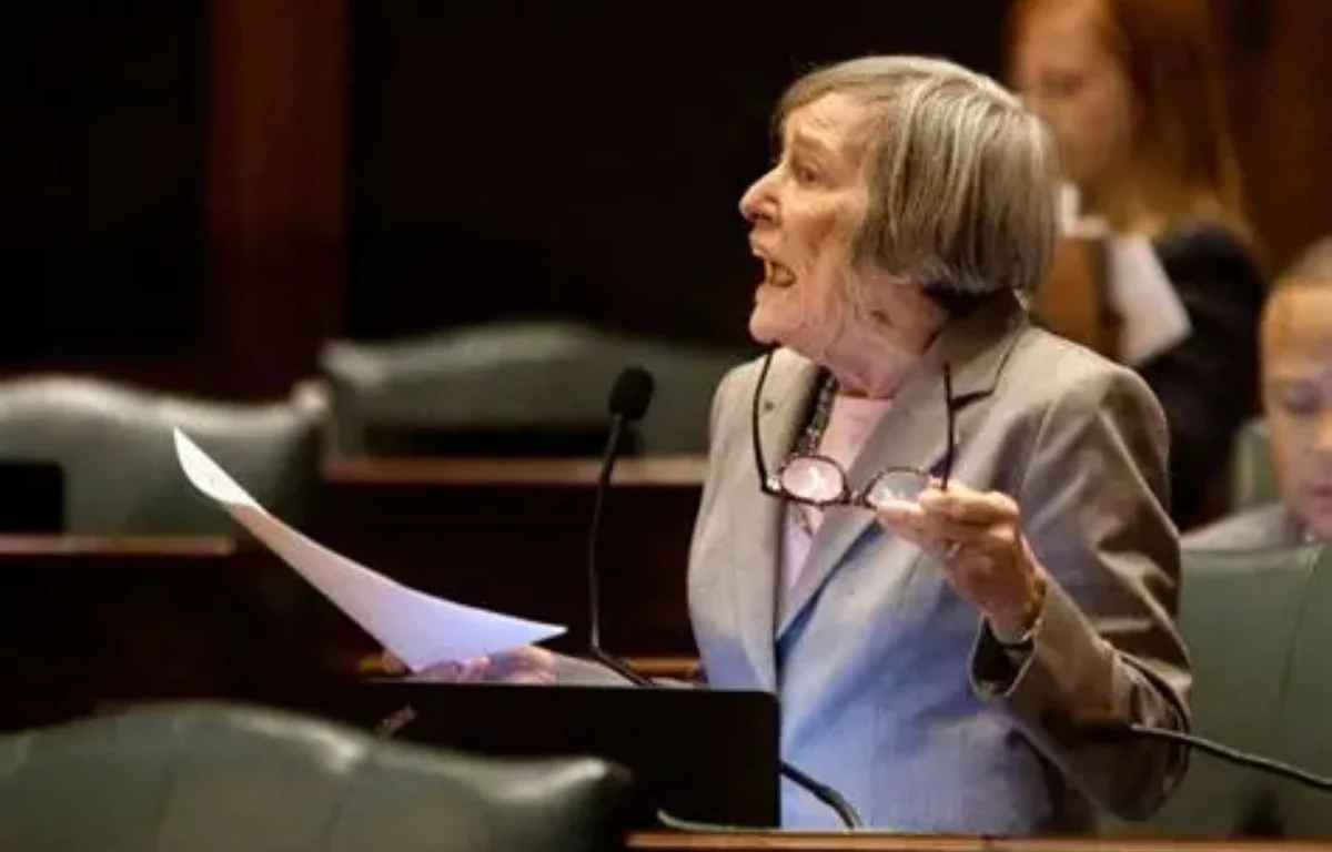 Older woman in a gray suit speaks at a podium with a microphone, holding papers and glasses.
