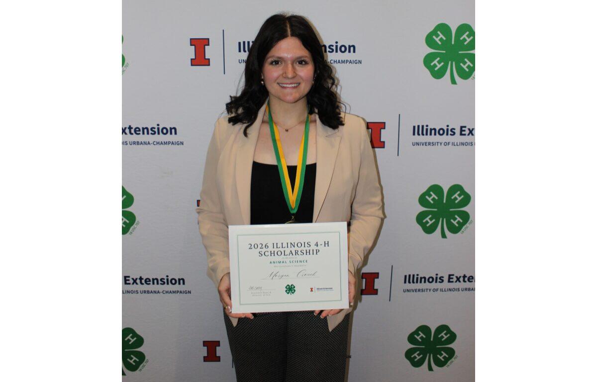 Smiling woman in a beige blazer holds a framed certificate that reads '2026 Illinois 4-H Scholarship' at a 4-H event backdrop.