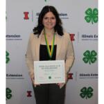 Smiling woman in a beige blazer holds a framed certificate that reads '2026 Illinois 4-H Scholarship' at a 4-H event backdrop.