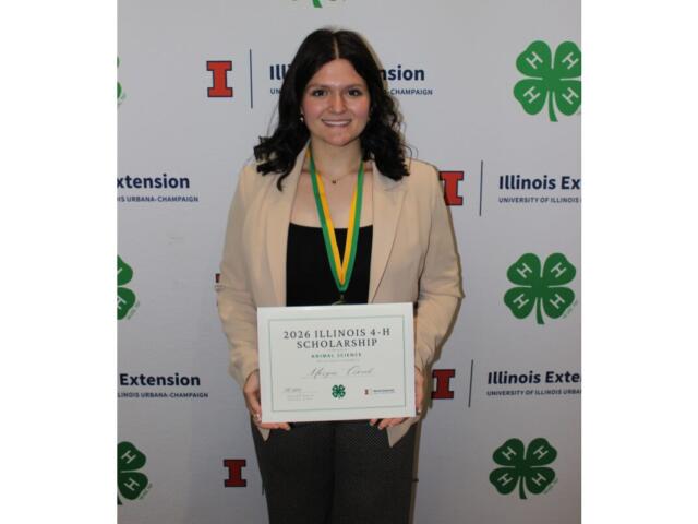 Smiling woman in a beige blazer holds a framed certificate that reads '2026 Illinois 4-H Scholarship' at a 4-H event backdrop.