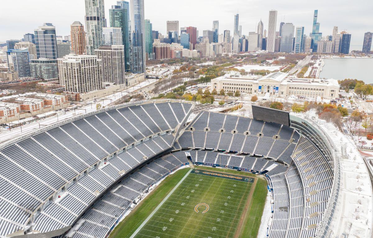 Aerial view of a large football stadium with snow, a green field, and a city skyline in the background.