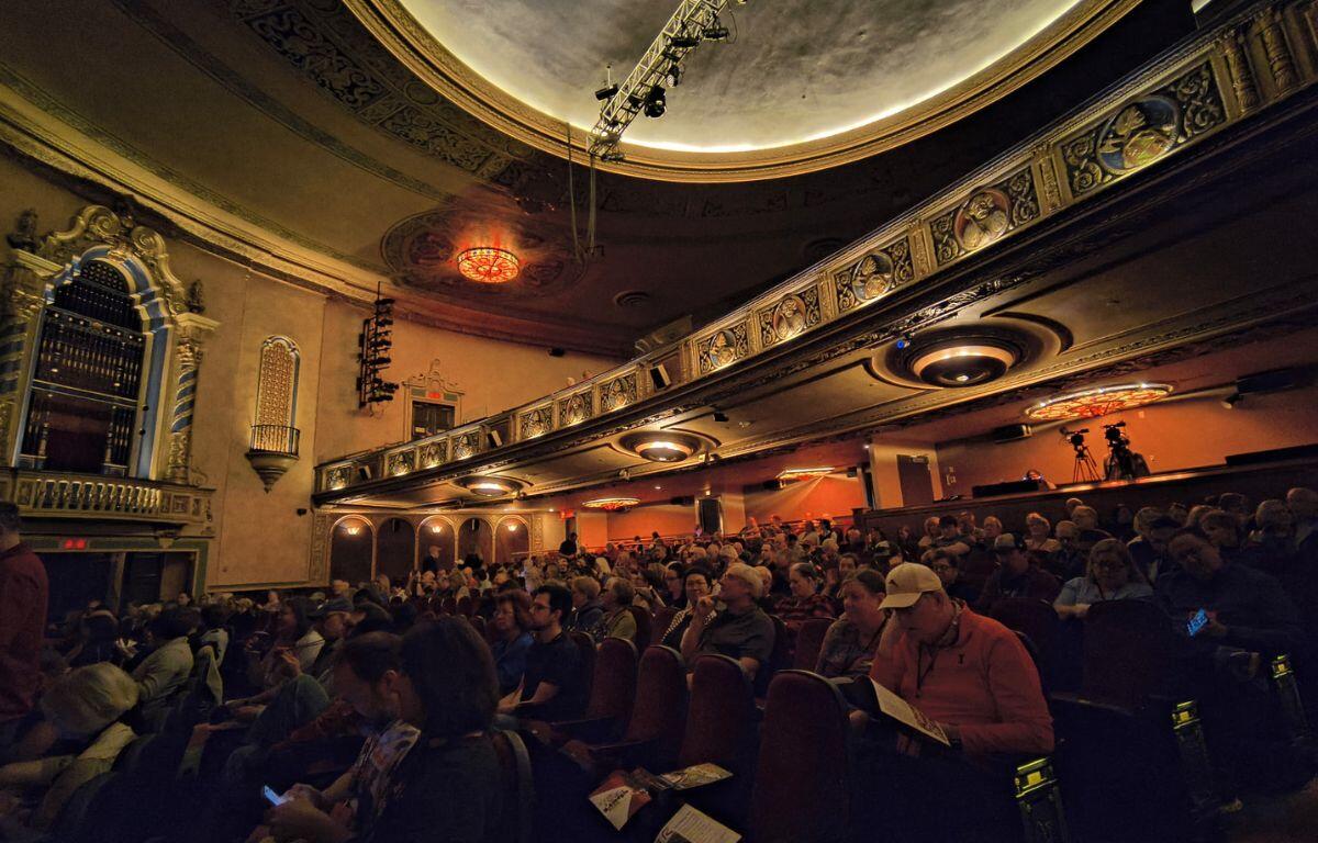 Interior of a historic theater with ornate balconies, warm lighting, and a full audience seated waiting for a performance