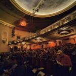 Interior of a historic theater with ornate balconies, warm lighting, and a full audience seated waiting for a performance