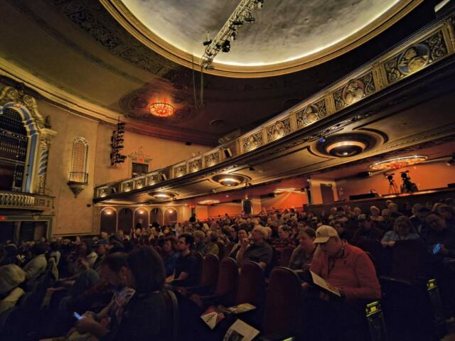 Interior of a historic theater with ornate balconies, warm lighting, and a full audience seated waiting for a performance