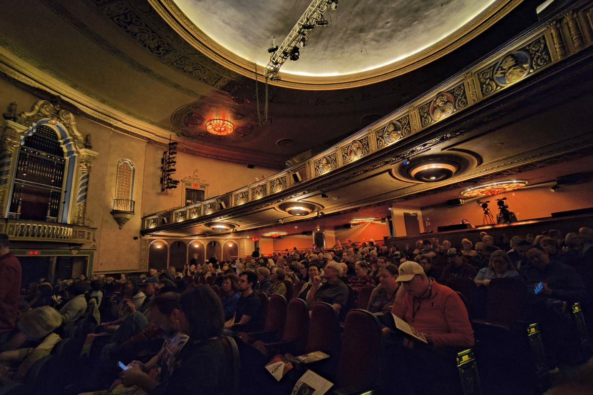Interior of a historic theater with ornate balconies, warm lighting, and a full audience seated waiting for a performance