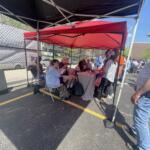 Group of people seated at a table under a red canopy at an outdoor event, near a chain-link fence on a sunny day