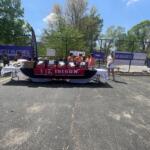Outdoor event scene with a long table draped in red and blue sports banners, volunteers in orange shirts behind it, and purple Best Buddies signage by a chain-link fence.