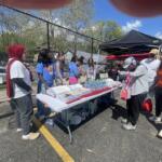 People face a long table of snacks and water bottles at an outdoor community event, with tents and a chain-link fence in the background.