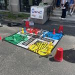 Outdoor street game setup with a large colorful board and pawns; cones and onlookers sit on steps nearby, sign reads 'Games'.