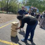 Person stacking wooden blocks into a tall Jenga-style tower at an outdoor event, with a colorful Kona Ice truck and crowd in the background.