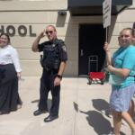 People stand outside a school entrance; a police officer salutes beside a smiling woman in a white blouse and another woman in a turquoise shirt.