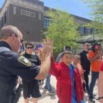 Police officer in a dark uniform raises a hand among a group of children in orange jackets outdoors, with a brick building in the background.