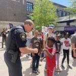 Police officer interacting with a group of smiling kids at an outdoor community event, with a Best Buddies Friendship Walk banner in the background.