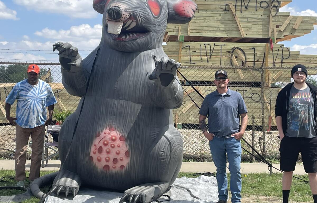 Large inflatable gray monster with red spots and claws, outdoors near a wooden structure; four people stand behind a chain-link fence.