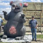 Large inflatable gray monster with red spots and claws, outdoors near a wooden structure; four people stand behind a chain-link fence.