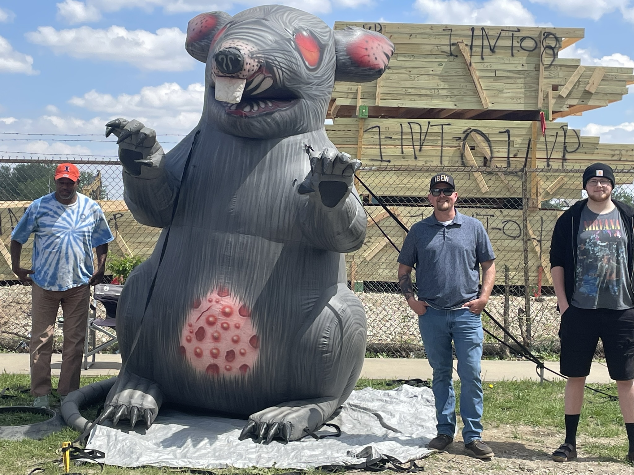 Large inflatable gray monster with red spots and claws, outdoors near a wooden structure; four people stand behind a chain-link fence.