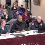 Three people sit at a front table with microphones and laptops; two police officers in uniform flank a woman in a green cardigan, guiding a meeting or hearing. Behind them, an audience sits in rows, with some attendees wearing masks, in a formal room labeled City of ... on the tablecloth.