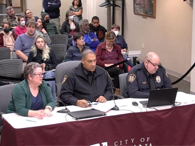 Three people sit at a front table with microphones and laptops; two police officers in uniform flank a woman in a green cardigan, guiding a meeting or hearing. Behind them, an audience sits in rows, with some attendees wearing masks, in a formal room labeled City of ... on the tablecloth.