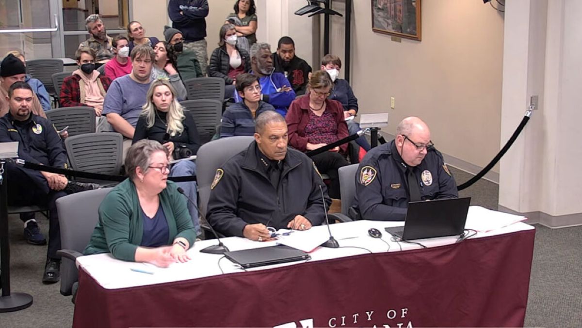 Three people sit at a front table with microphones and laptops; two police officers in uniform flank a woman in a green cardigan, guiding a meeting or hearing. Behind them, an audience sits in rows, with some attendees wearing masks, in a formal room labeled City of ... on the tablecloth.