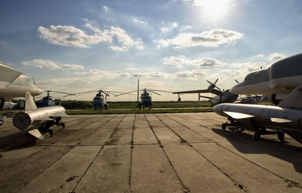 Several aircraft are parked on a concrete airstrip, with a row of blue helicopters in the distance under a bright, sunny sky.