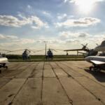 Several aircraft are parked on a concrete airstrip, with a row of blue helicopters in the distance under a bright, sunny sky.