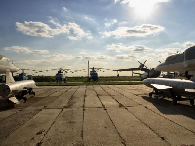 Several aircraft are parked on a concrete airstrip, with a row of blue helicopters in the distance under a bright, sunny sky.