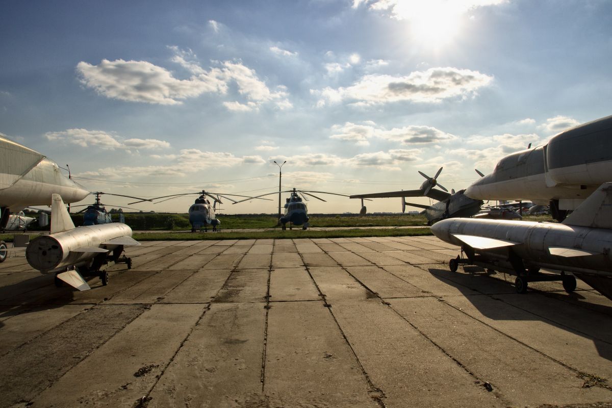 Several aircraft are parked on a concrete airstrip, with a row of blue helicopters in the distance under a bright, sunny sky.