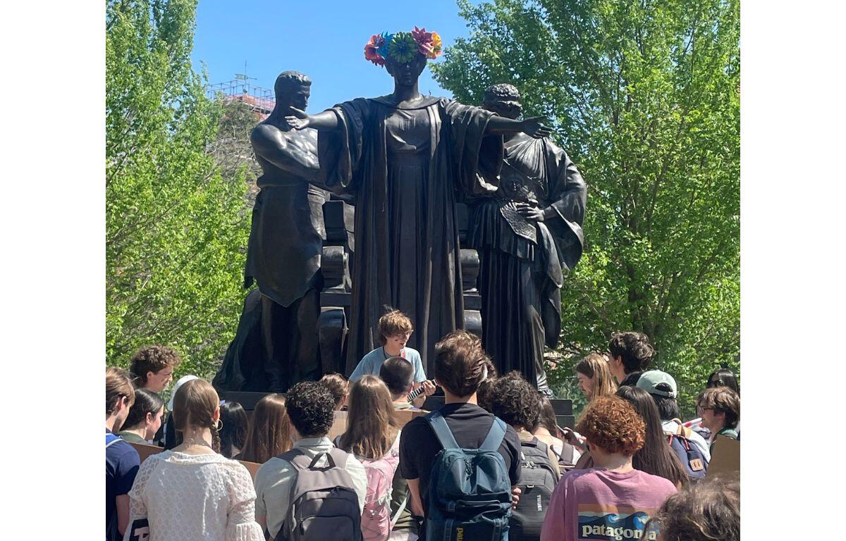 Group of students gathered around a large dark sculpture of three figures in a park; the central figure wears a colorful flower crown.