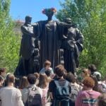 Group of students gathered around a large dark sculpture of three figures in a park; the central figure wears a colorful flower crown.
