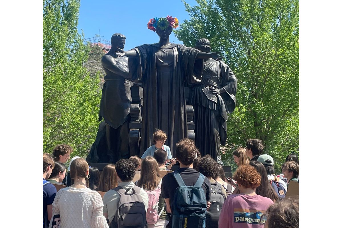 Group of students gathered around a large dark sculpture of three figures in a park; the central figure wears a colorful flower crown.