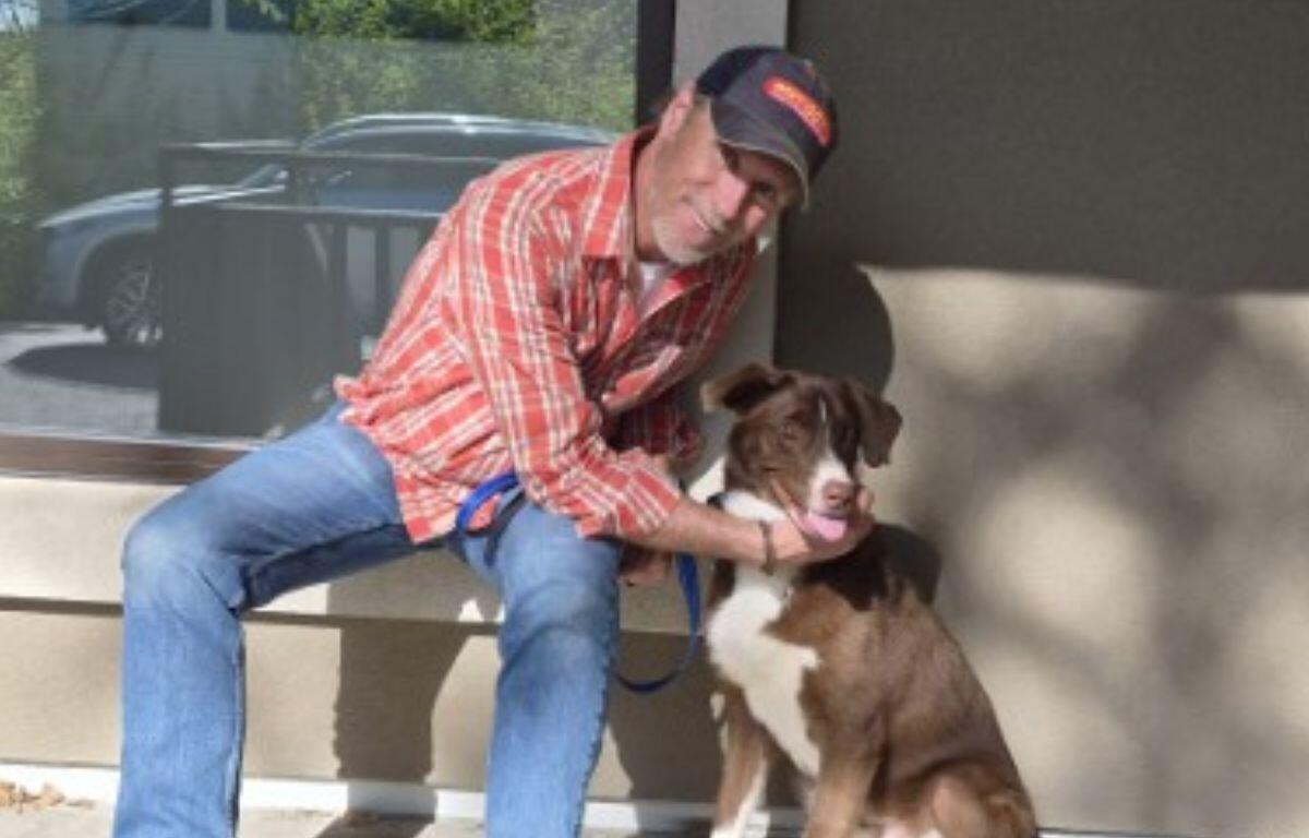 An older man in a red plaid shirt and cap sits on a bench outside, holding a brown and white dog on a leash and sharing a moment together.