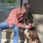 An older man in a red plaid shirt and cap sits on a bench outside, holding a brown and white dog on a leash and sharing a moment together.