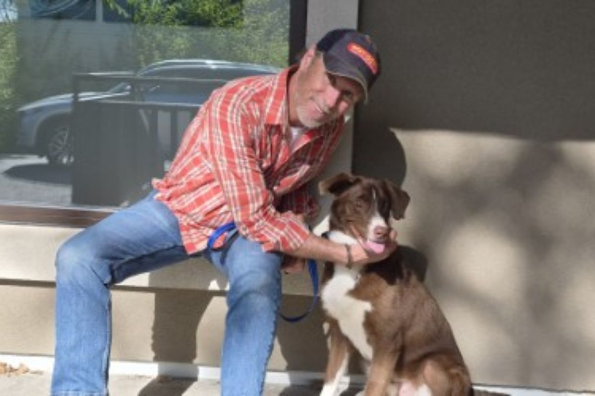 An older man in a red plaid shirt and cap sits on a bench outside, holding a brown and white dog on a leash and sharing a moment together.