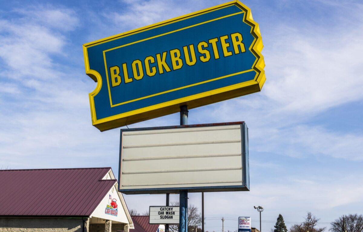Large blue-and-yellow Blockbuster sign atop a pole with an empty white marquee beneath, clear sky in the background.