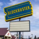 Large blue-and-yellow Blockbuster sign atop a pole with an empty white marquee beneath, clear sky in the background.