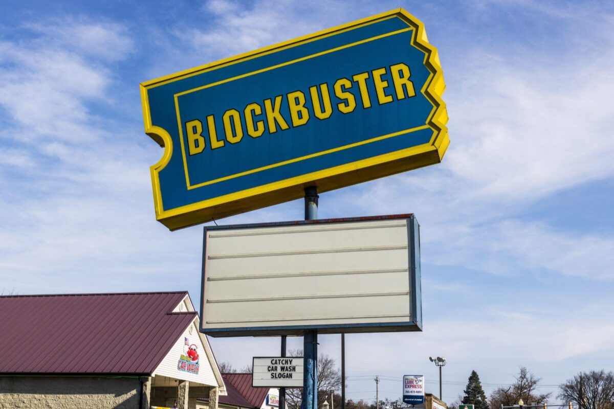 Large blue-and-yellow Blockbuster sign atop a pole with an empty white marquee beneath, clear sky in the background.