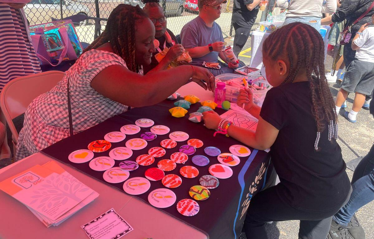 Woman and young girl craft colorful round badges at an outdoor community table.