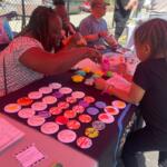 Woman and young girl craft colorful round badges at an outdoor community table.