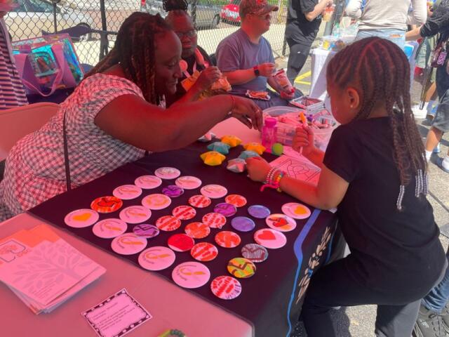 Woman and young girl craft colorful round badges at an outdoor community table.