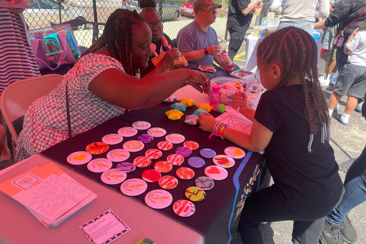 Woman and young girl craft colorful round badges at an outdoor community table.