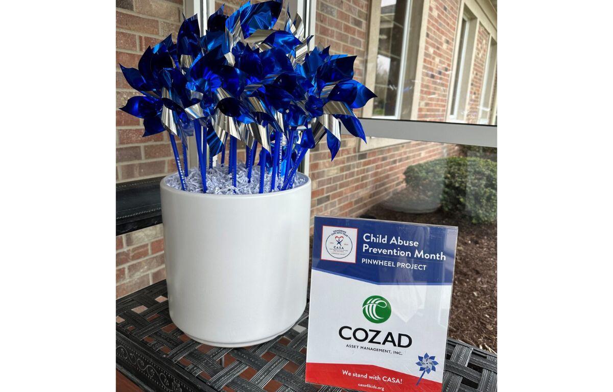 Blue metallic pinwheels arranged in a white pot for Child Abuse Prevention Month, placed on a patio beside a brick wall with a CASA/COZAD sign nearby.