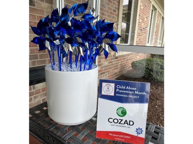 Blue metallic pinwheels arranged in a white pot for Child Abuse Prevention Month, placed on a patio beside a brick wall with a CASA/COZAD sign nearby.