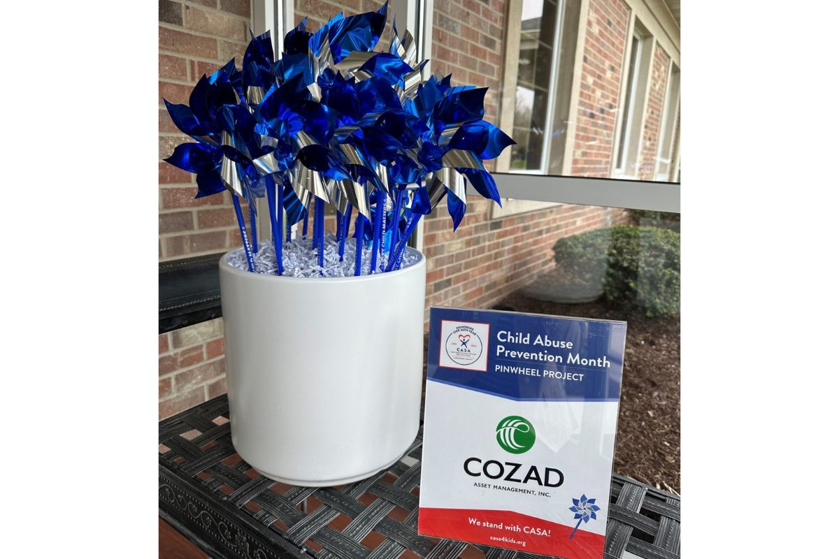 Blue metallic pinwheels arranged in a white pot for Child Abuse Prevention Month, placed on a patio beside a brick wall with a CASA/COZAD sign nearby.