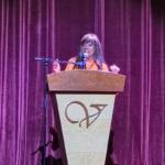 Woman delivering a speech at a wooden podium on a stage with burgundy curtains behind her.