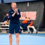 Male coach in a navy Illinois uniform on an indoor blue court, clapping his hands.