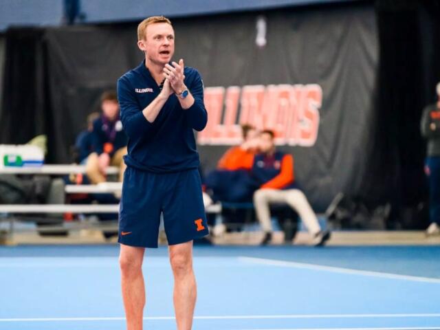 Male coach in a navy Illinois uniform on an indoor blue court, clapping his hands.