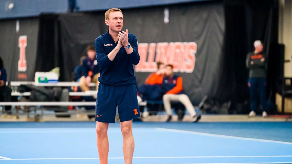Male coach in a navy Illinois uniform on an indoor blue court, clapping his hands.