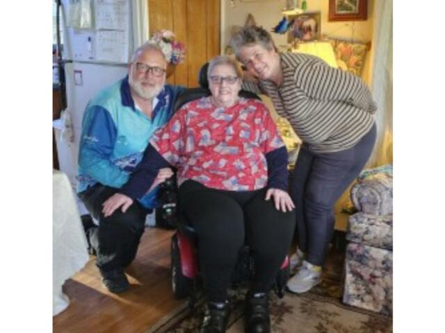 Three people posing in a living room: an elderly woman in a wheelchair flanked by two smiling companions.