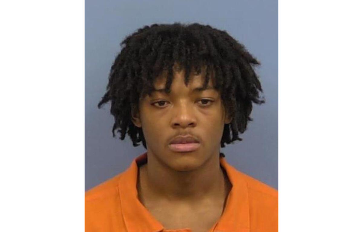 Mugshot of a young man with short dreadlocks wearing an orange jail uniform, facing the camera against a gray background.